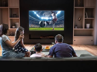 A family of 4 sat on the sofa facing their TV watching a man kick a ball